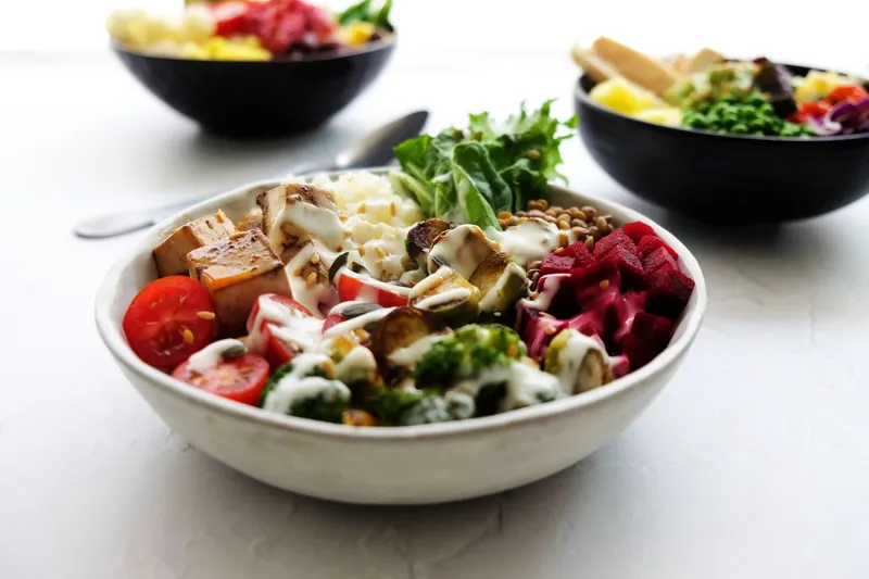 Weiße Schüssel mit Tofuwürfeln, Rote Bete, Tomaten, Rucola und Joghurtsauce, dahinter zwei weitere Bowls auf hellem Tisch